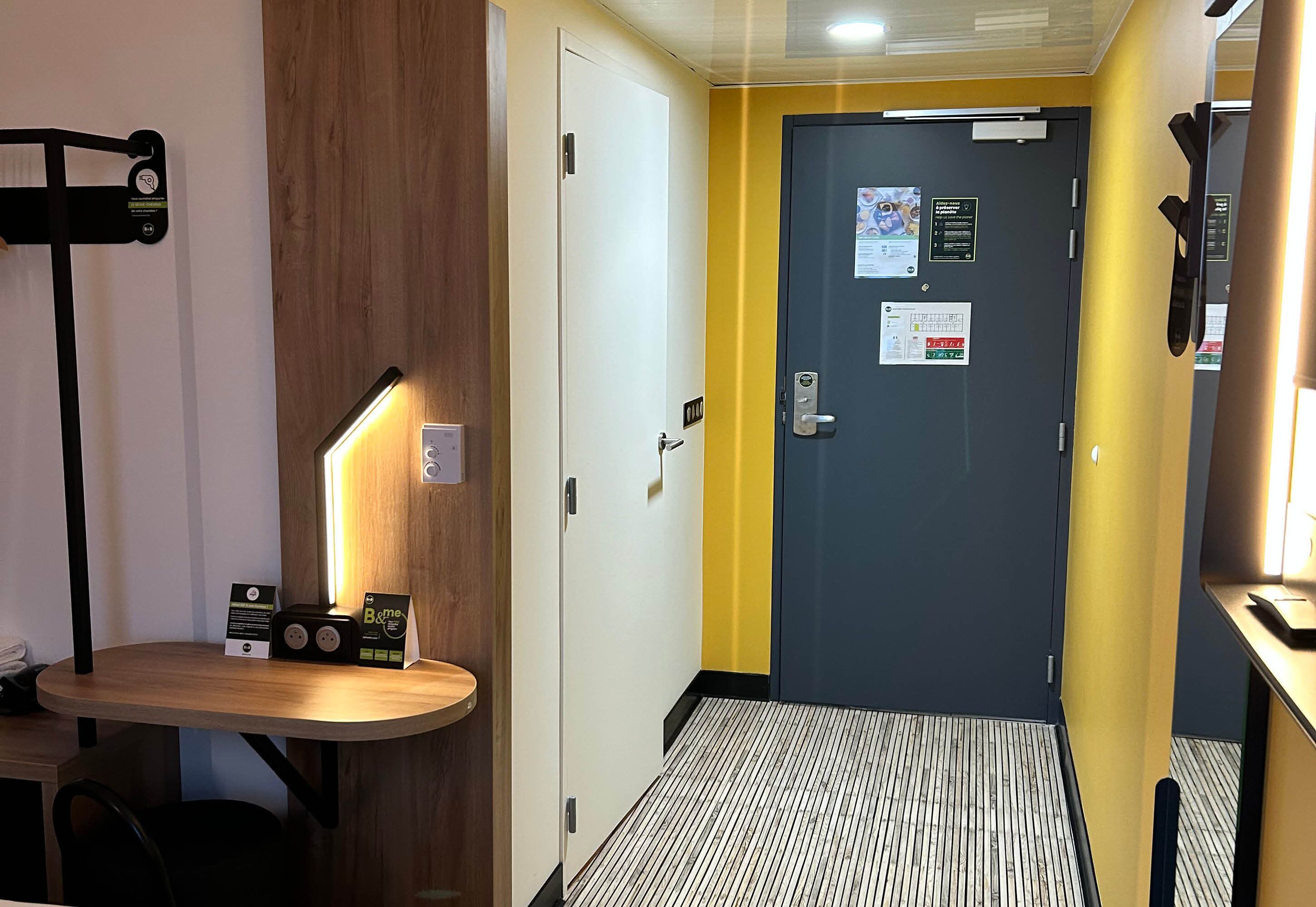 Een hotelkamer met een houten bureau, een staande lamp, een deur met veiligheidsslot en een vloer met patroon.