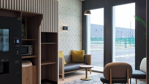 Moderni hotellihuone, jossa on sisäänrakennettu mikroaaltouuni, oleskelualue sohvineen ja nojatuoleineen, pieni pöytä sekä suuret ikkunat, joista on näkymä ulos.