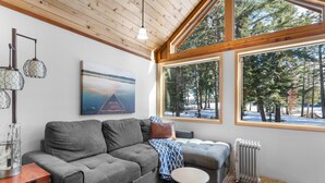 Una sala de estar acogedora con un sofá gris, una mesa de centro y un radiador. Amplias ventanas ofrecen vistas a árboles y un paisaje nevado.