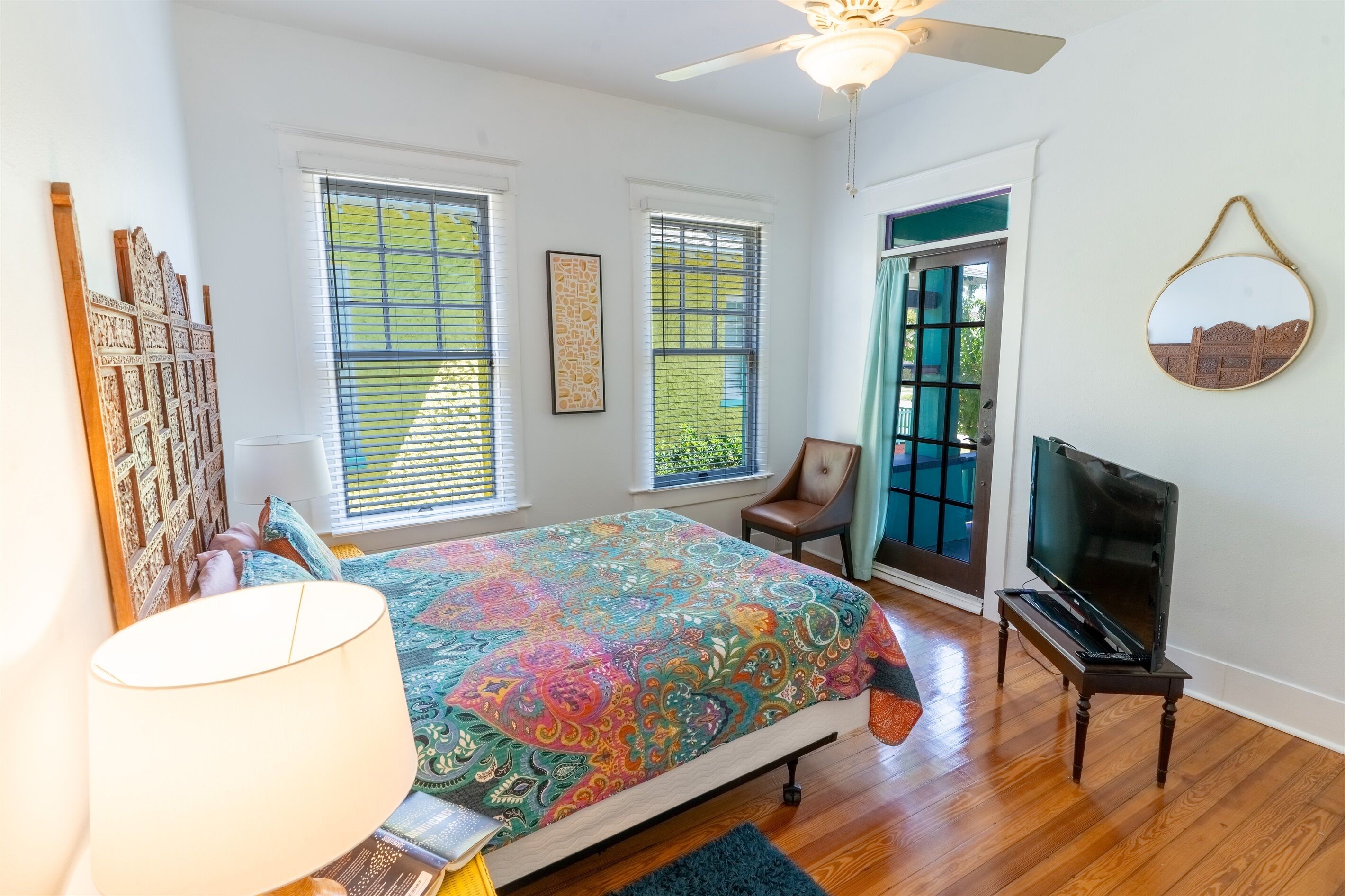 A bedroom with a bed, a TV, a ceiling fan, and large windows.
