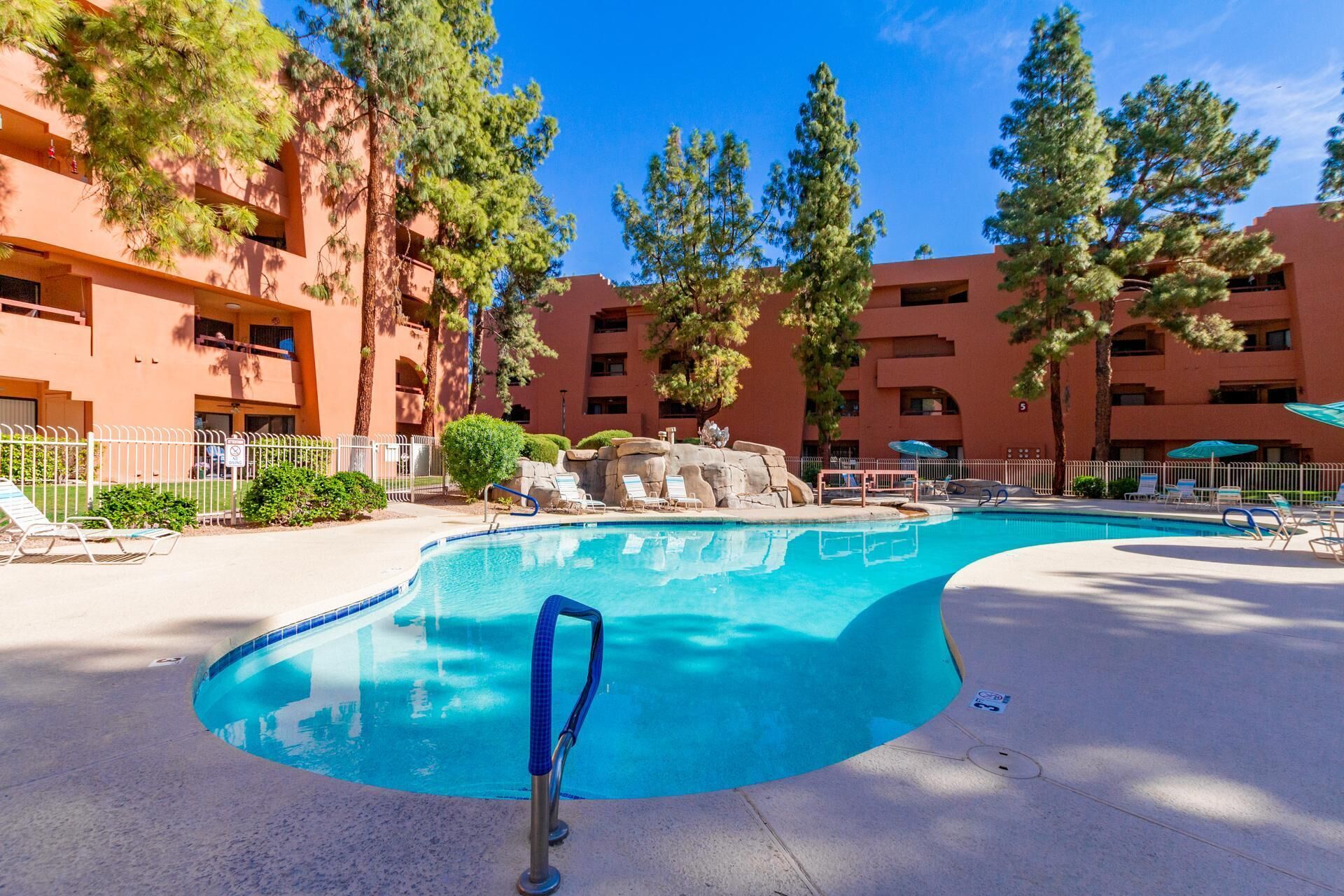 Outdoor pool area with curved blue handrails, lounge chairs, umbrellas, and tall pine trees surrounding terracotta buildings.