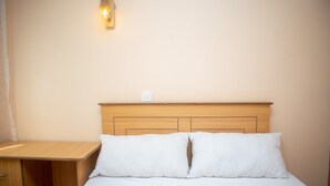 A neatly made bed with two pillows, a wooden nightstand with drawers, and a wall-mounted light fixture. - Korongoro Hotel Chemolingot (Chemolingot)