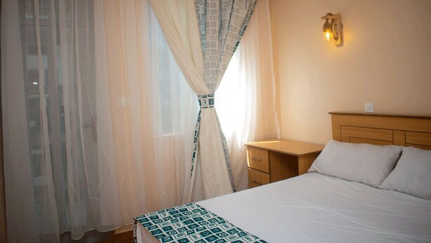 A hotel room with a bed, a nightstand, a wall-mounted light, and sheer curtains with a patterned tie-back. - Korongoro Hotel Chemolingot (Chemolingot)