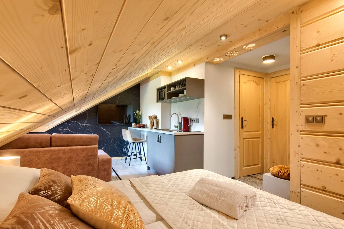 A bedroom with a bed, a seating area, a kitchenette, and wooden cabinets.
