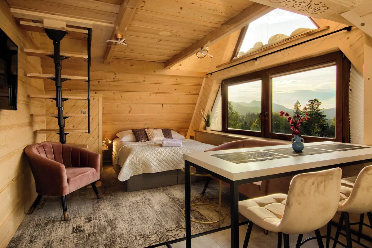 A cozy bedroom with a bed, a desk, and chairs, a window with a view of mountains, and a wooden ceiling.