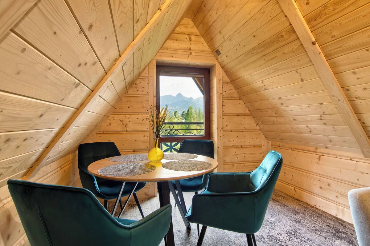 A wooden interior with a round table and four chairs, a window with a mountain view, and a door with a decorative design.