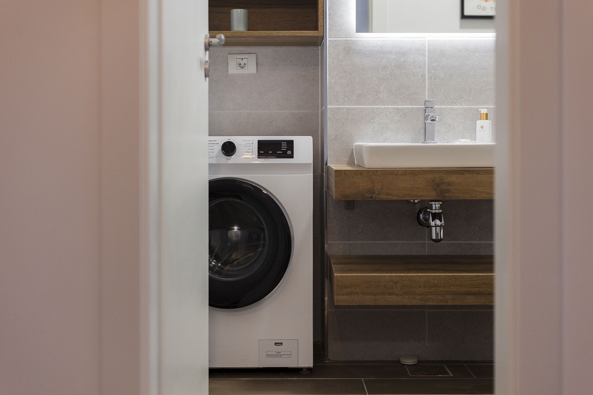 Un lavadero pequeño con lavadora de carga frontal, un lavabo y un estante de madera.