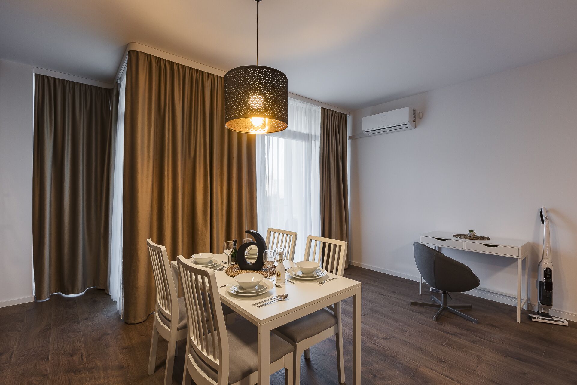 Un comedor con una mesa lista para cuatro personas, una silla moderna, un escritorio con una computadora y un equipo de aire acondicionado en la pared.