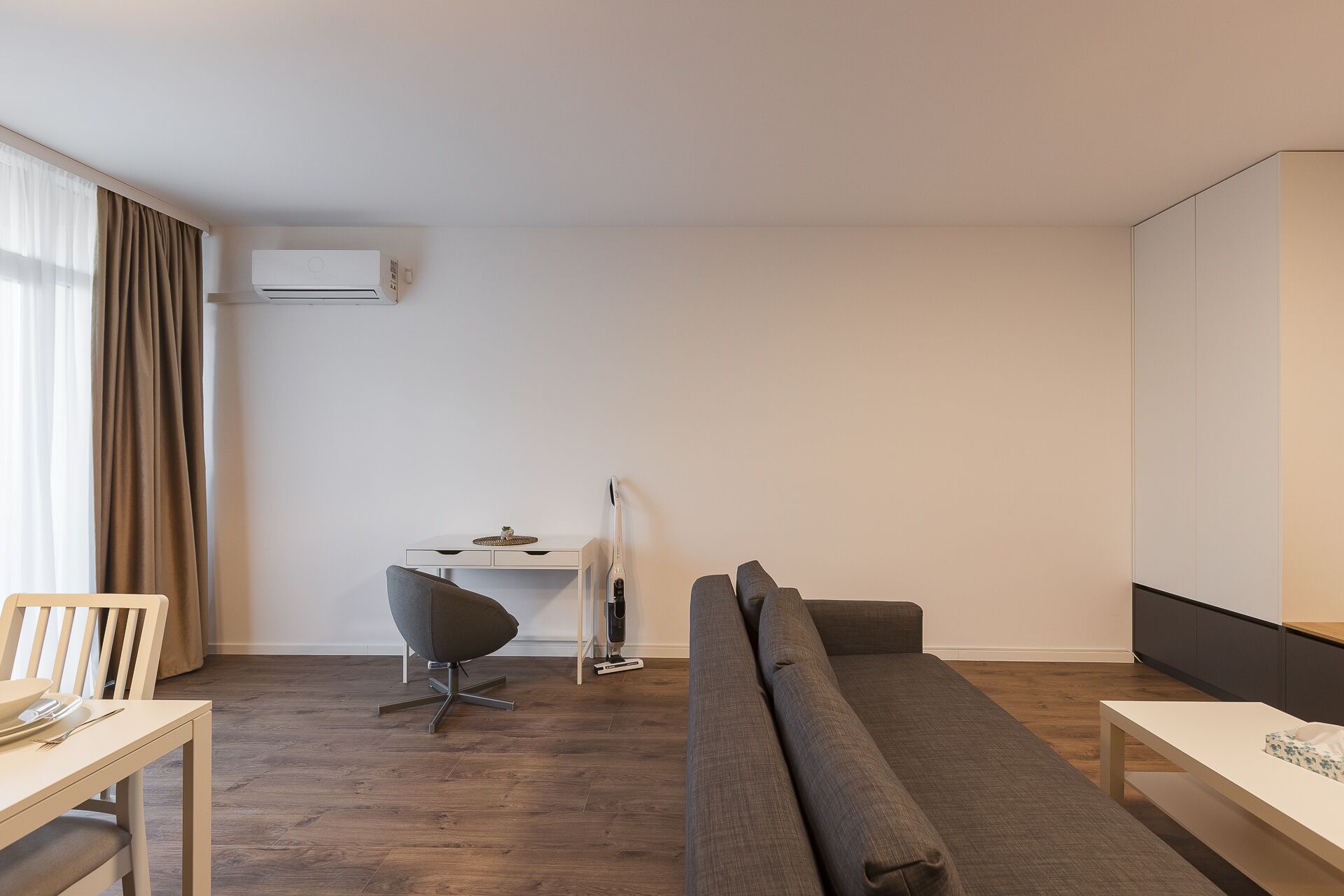 A modern hotel room with a wooden floor, a sofa, a dining table with chairs, a desk with a chair, and a television.