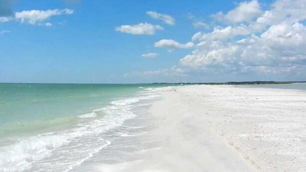 Une plage à l’eau turquoise et limpide, bordée de sable blanc, sous un ciel partiellement nuageux.