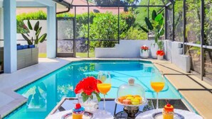 Un espace aménagé au bord de la piscine, doté d’une structure en verre, d’une table avec des boissons et des pâtisseries, et de plantes en pot.