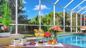Desayuno preparado junto a una piscina, con una mesa, tazas de café, fruta y flores.