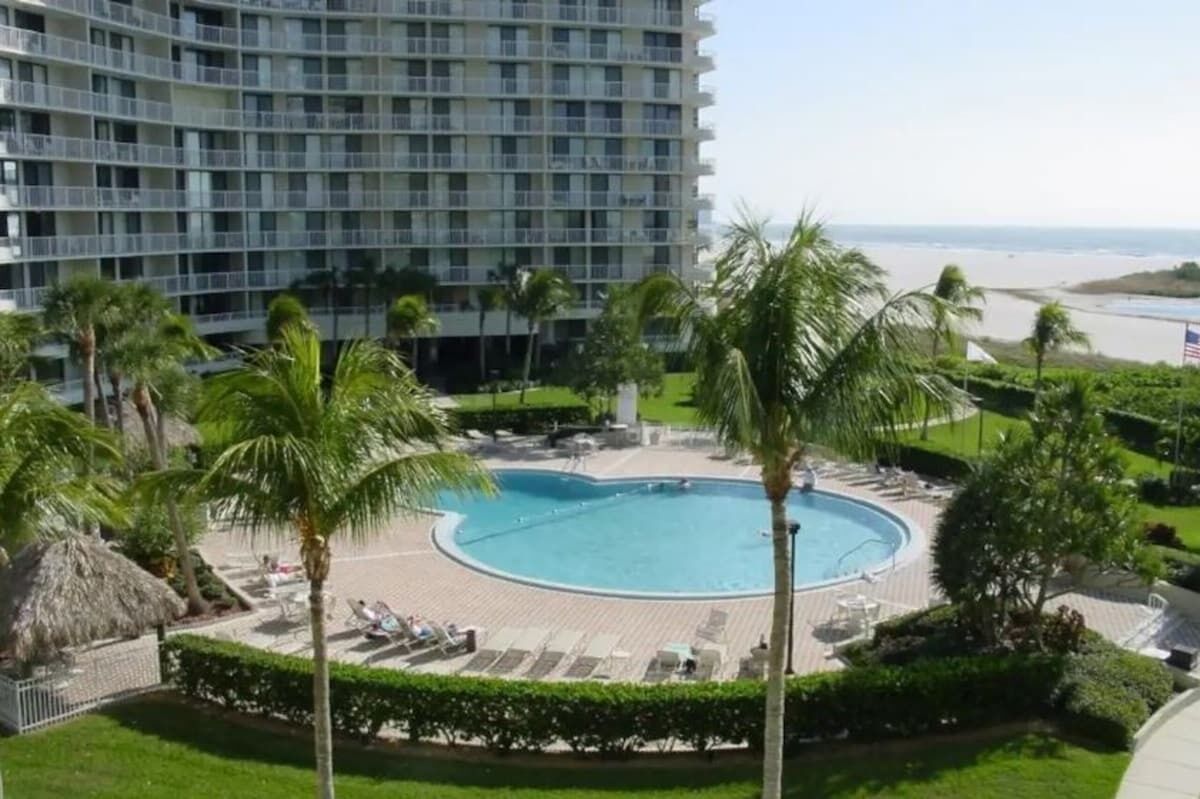 Un hôtel doté d’une grande piscine entourée de palmiers et de chaises longues, avec vue sur une plage.