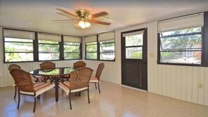 A dining area with a round table and chairs, a ceiling fan, and large windows with blinds.