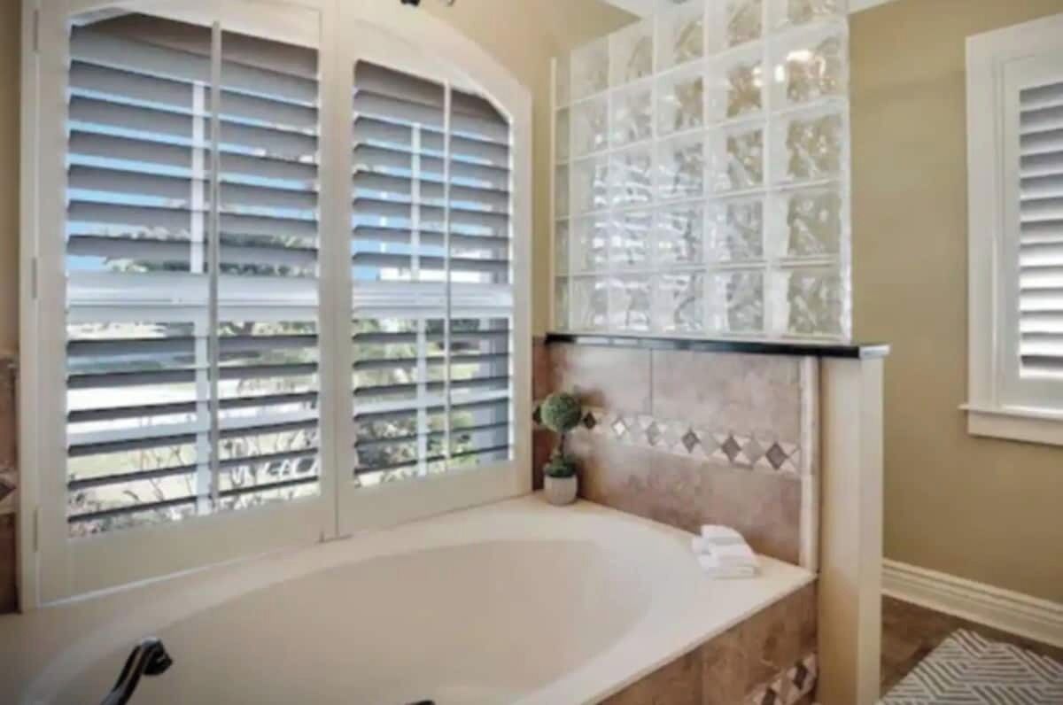 Ein Badezimmer mit Badewanne, einem Fenster mit Jalousien und einer gefliesten Rückwand.