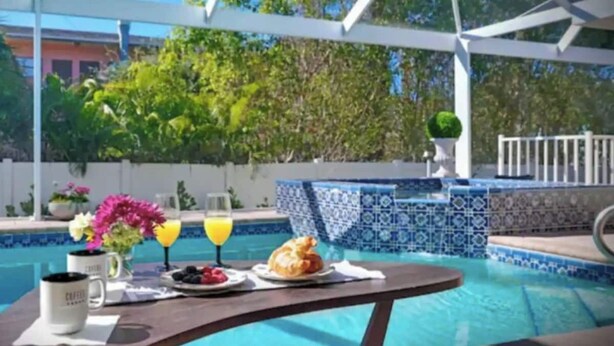 Une table placée au bord de la piscine, avec des aliments pour le petit-déjeuner, un verre de jus d’orange et un vase avec des fleurs.