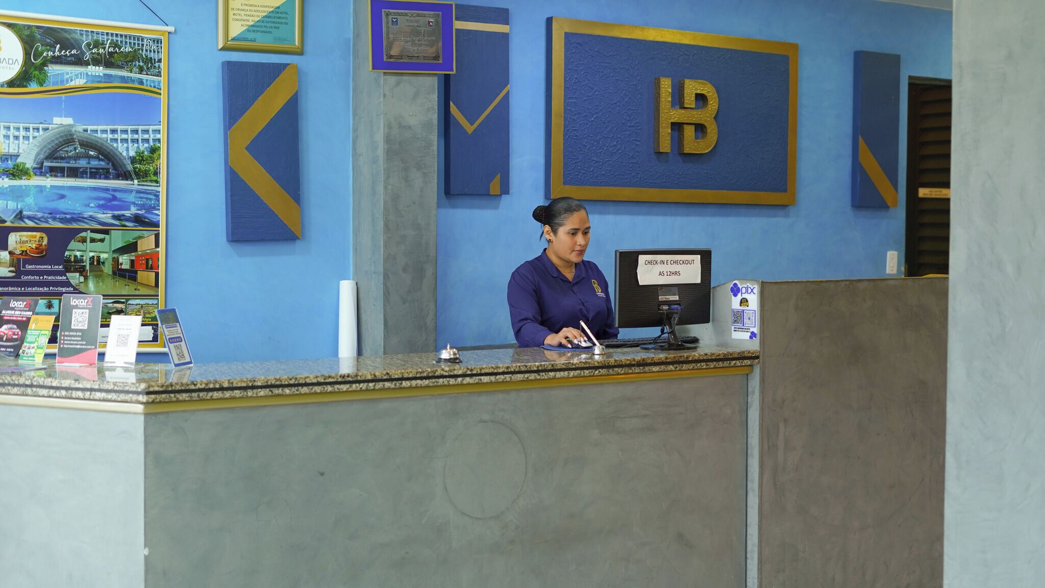 Un mostrador de recepción de hotel con una decoración azul y dorada, un certificado enmarcado en la pared y un cartel que indica los horarios de registro de entrada y salida.