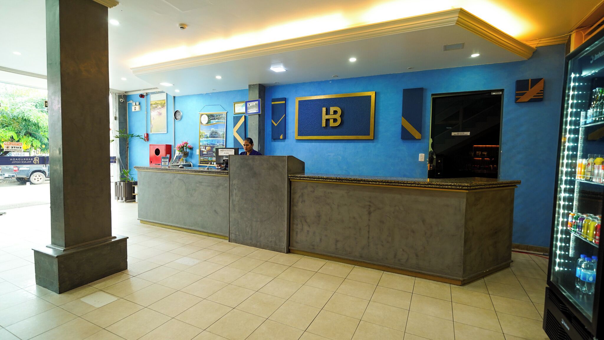 Un área de recepción con pared azul, mostrador de recepción, refrigerador con bebidas y ventana con vista a la calle.