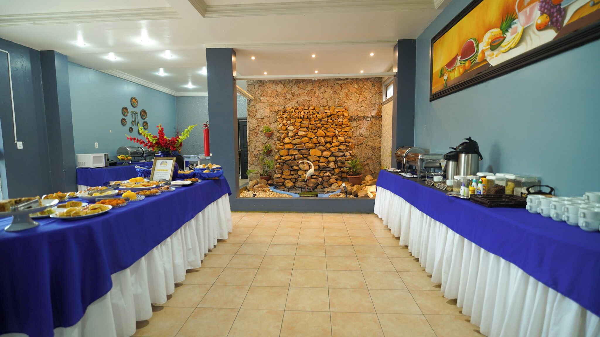 A buffet setup with food items on tables, a stone wall, and a painting on the wall.