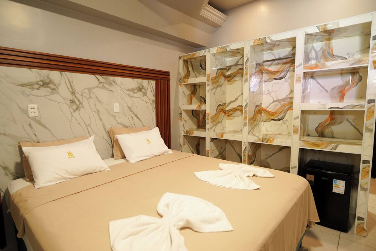 Hotelkamer met een marmeren hoofdeinde, een bed met twee kussens, een minibar en een inbouwplank met decoratieve objecten.