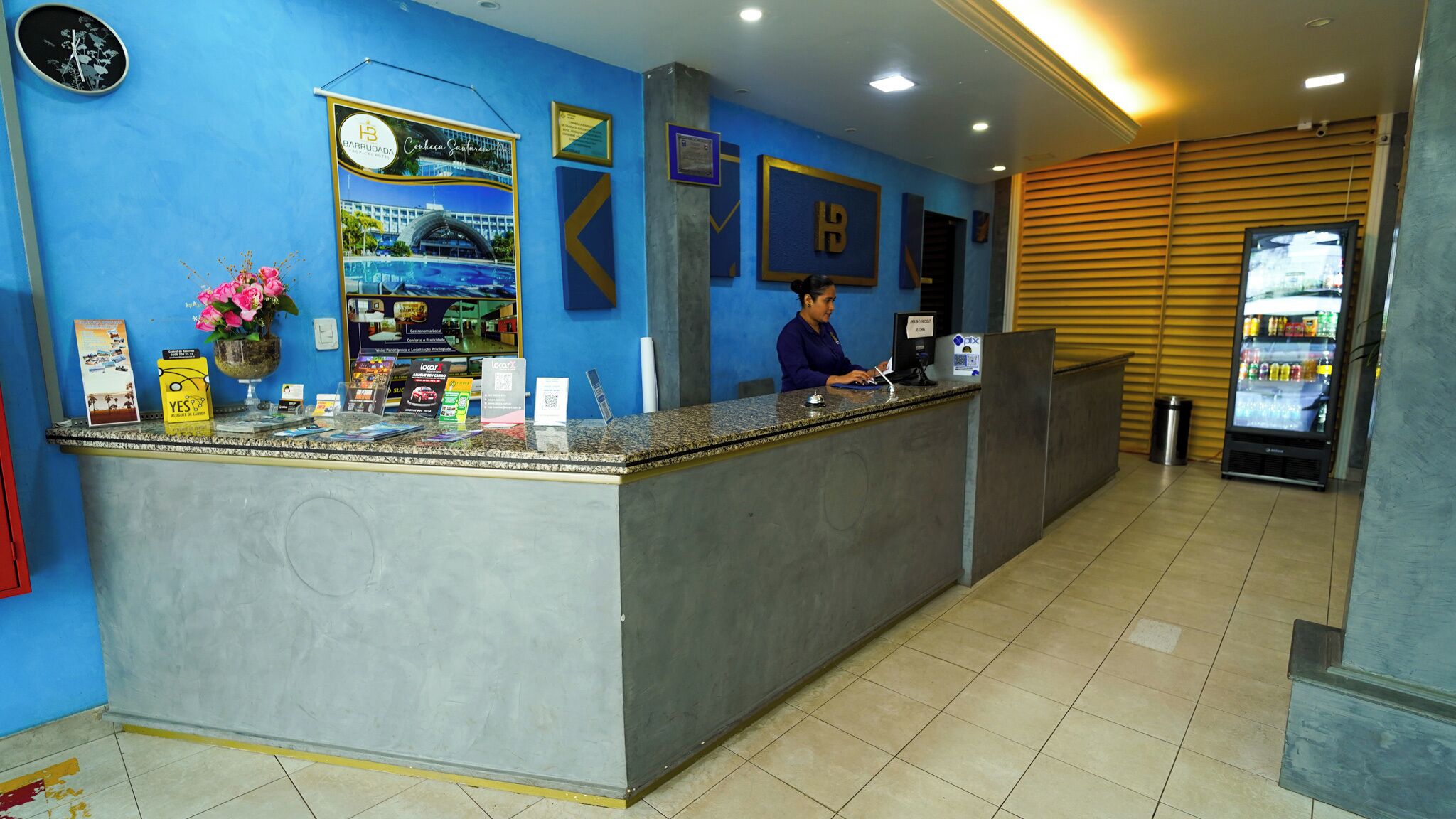 A hotel reception area with a blue wall, a receptionist, a reception desk, a clock, and a refrigerator.