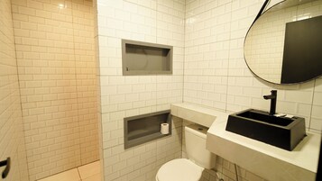 Un baño con azulejos blancos tipo metro, un inodoro, un lavabo con espejo y un cubo de basura negro.