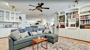 A living room with a sectional sofa, a wooden coffee table, and a patterned rug. There are bookshelves filled with books and a ceiling fan.