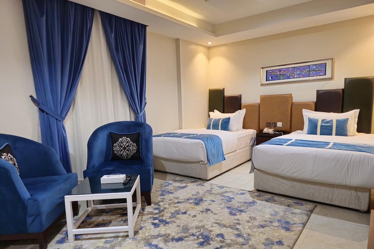 Habitación de hotel con dos camas, un sillón azul, una mesita de noche blanca y una alfombra con estampado azul y blanco.