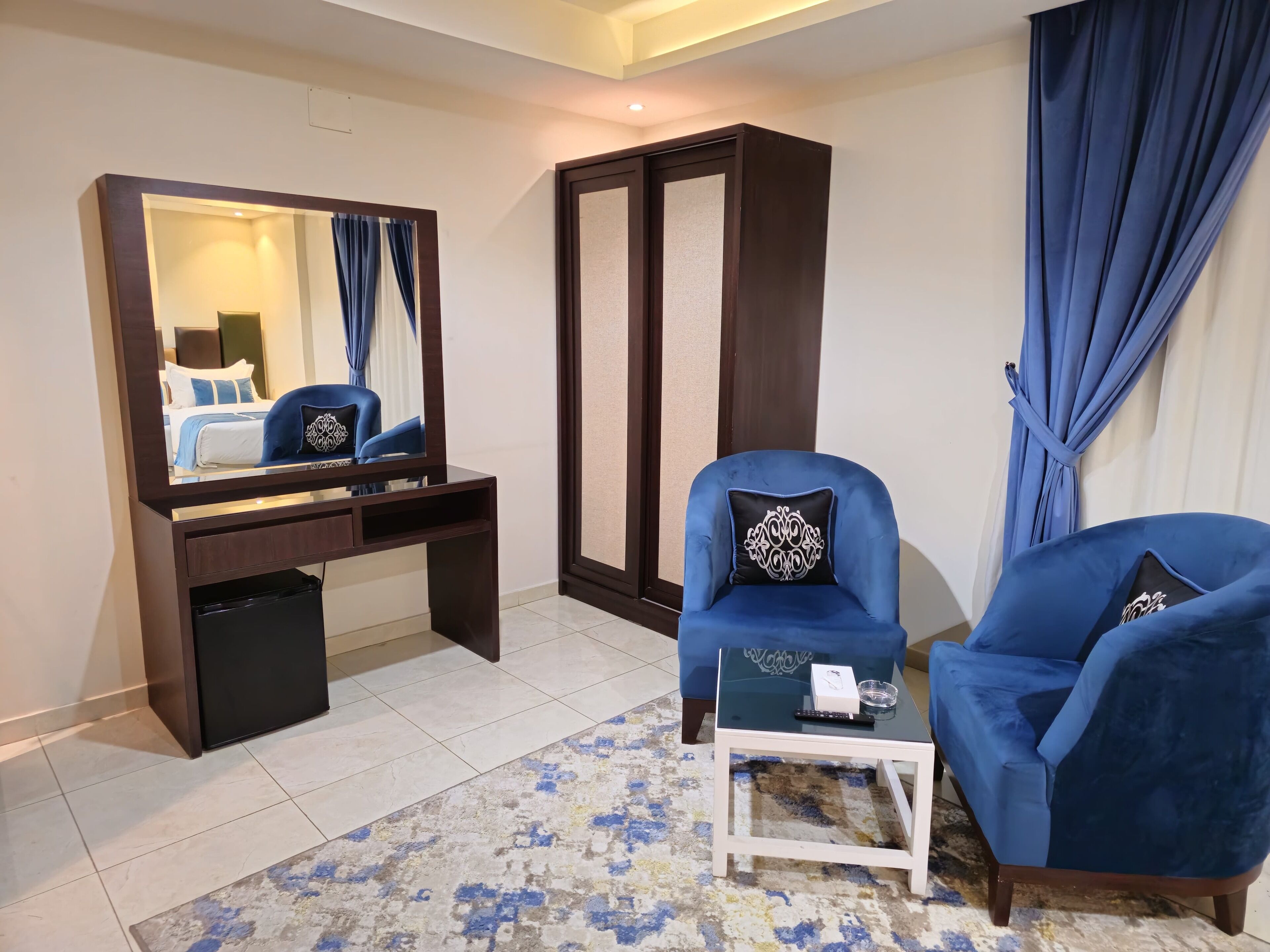 A hotel room with a large mirror, a dark wooden desk, two blue armchairs, a small white side table, and a patterned rug.