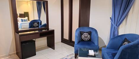 A hotel room with a large mirror, a dark wooden desk, two blue armchairs, a small white side table, and a patterned rug.