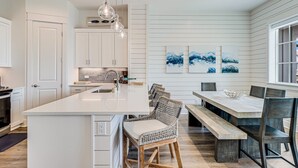 A modern kitchen with white cabinetry, a large island, and a dining area with wooden benches and chairs. - Three-story townhouse with a balcony, two living areas, & a washer/dryer (Port Aransas)
