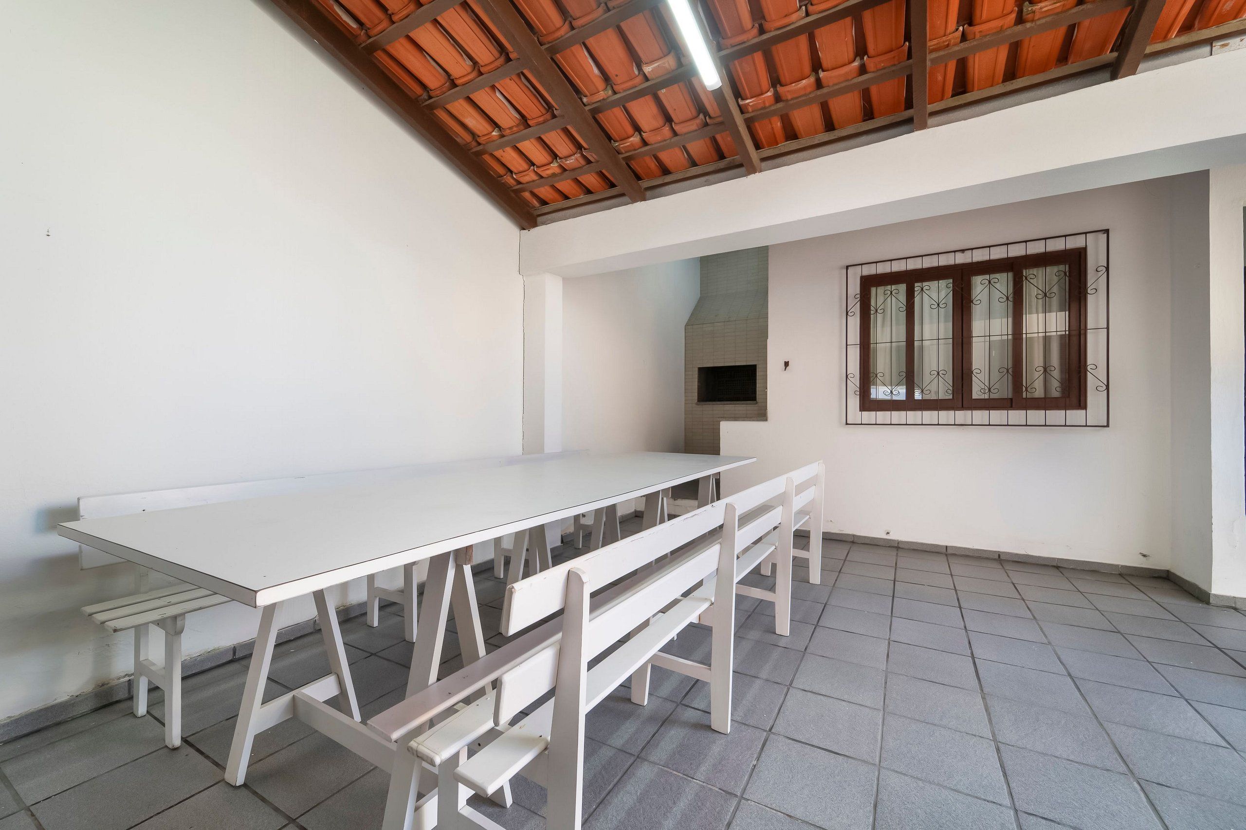 Um quarto com uma mesa branca comprida e bancos, piso e teto azulejados, e uma janela com grades.