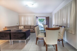 A living room with a sofa, chairs, a dining table, and a view through a window. - 914 - Casa 3 dormitórios para 8 pessoas (Bombinhas)