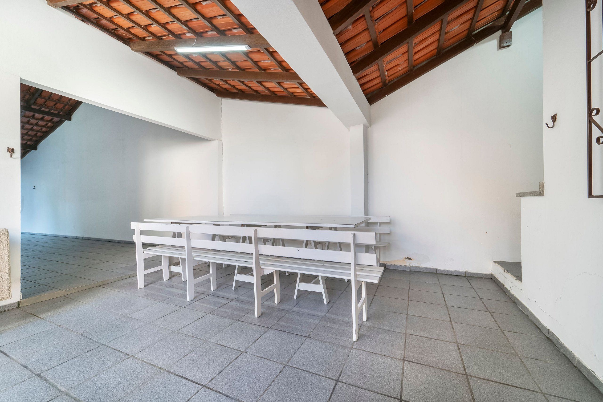 Um quarto com soalho em azulejos, paredes brancas, teto de madeira e uma mesa branca com bancos.