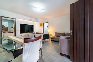 A modern living room with a glass dining table, a flat-screen TV, a sofa, and a wooden wardrobe.