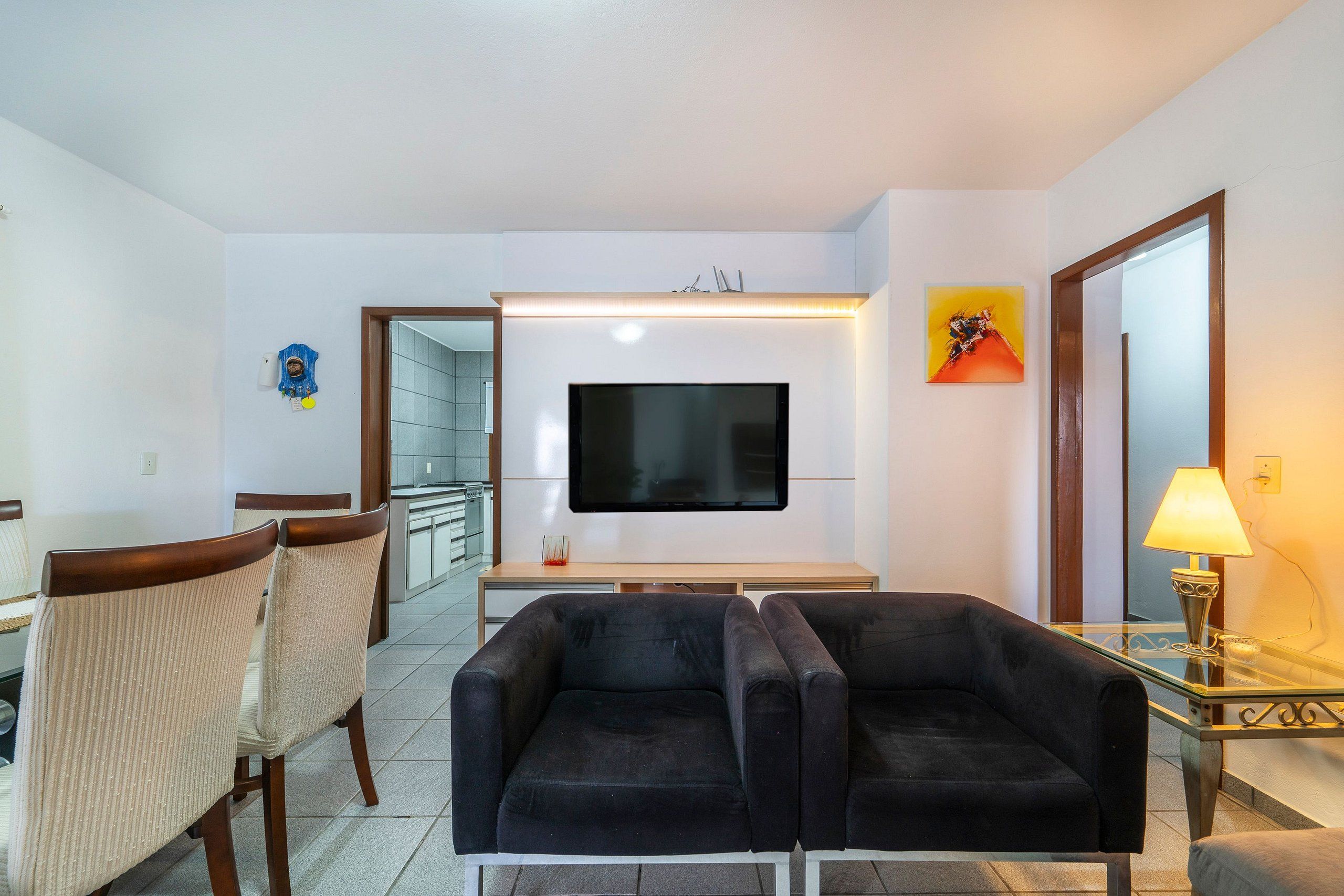 Uma sala de estar com televisão de tela plana, dois sofás pretos, uma área de refeições com cadeiras, uma mesa de centro de vidro e um abajur.