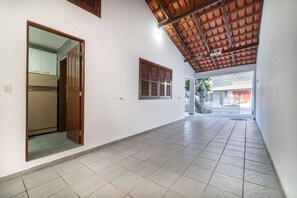 A corridor with tiled flooring, a wooden door, a window with shutters, and a view of a building outside. - 914 - Casa 3 dormitórios para 8 pessoas (Bombinhas)