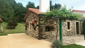 Een stenen huis met pannendak, schoorsteen en een kleine tuin met potplanten en een tuinslang.
