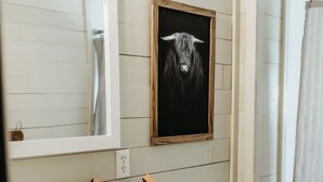 Un baño con un lavamanos blanco, un espejo, un cuadro enmarcado de una vaca, un inodoro y una cortina de ducha.