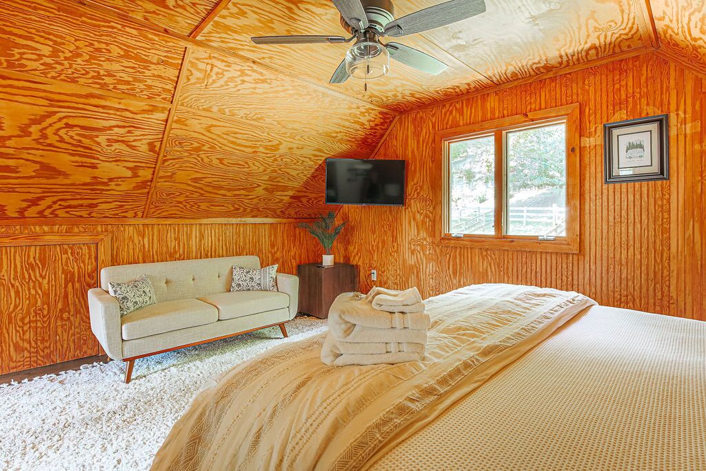 A cozy room with wooden walls, a bed with a patterned bedspread, a white sofa with cushions, a television on the wall, and a window with a view of greenery.