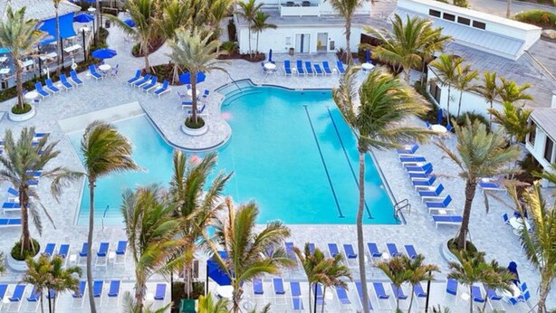 Luchtbeeld van een resort met een centraal gelegen zwembad, omringd door ligbedden en palmbomen.