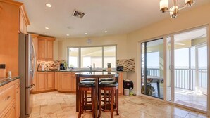 A kitchen with wooden cabinets, a stainless steel refrigerator, a central island with stools, a large window, and a sliding glass door leading to a balcony. - Beautiful Open-Water Property (Big Pine Key)