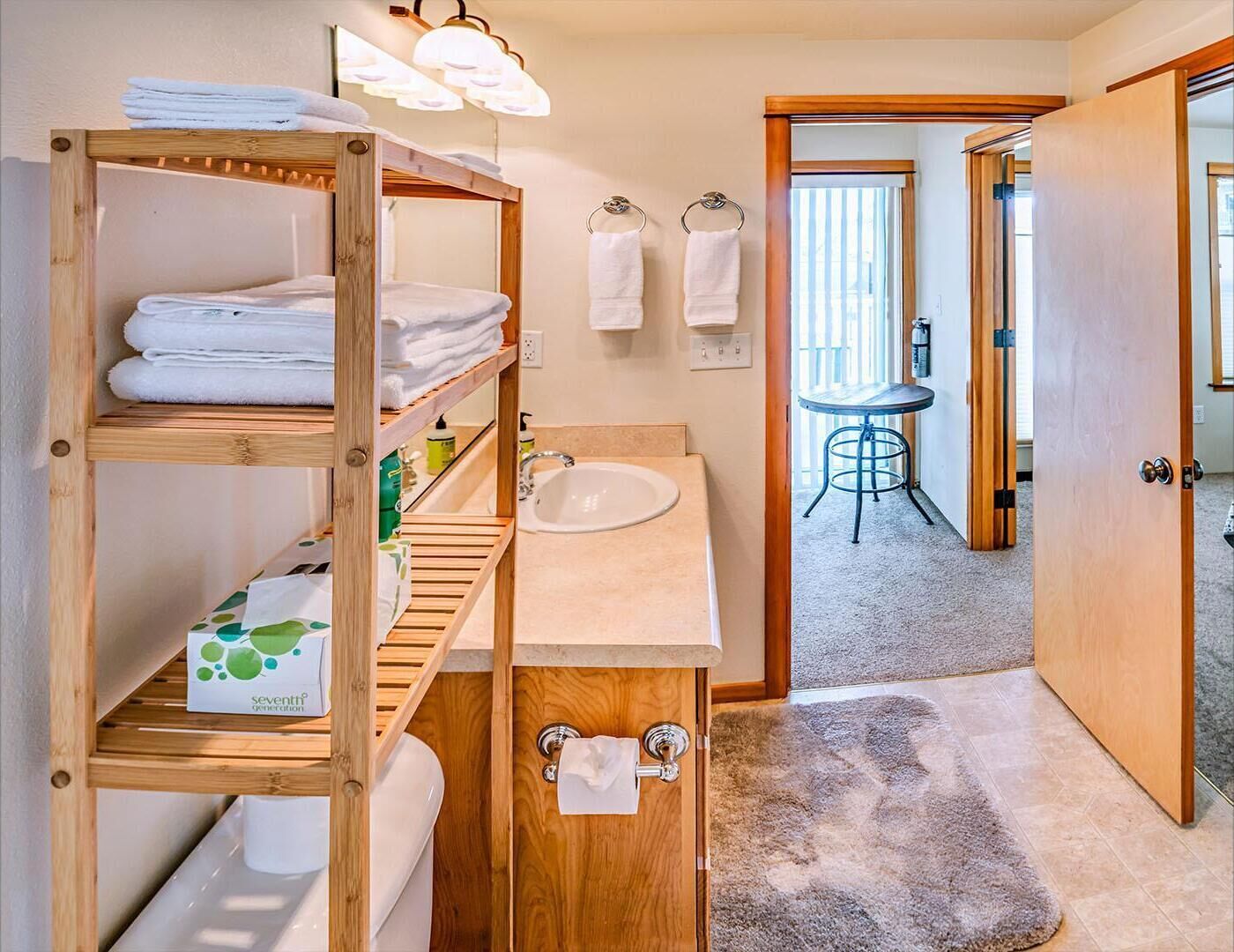 A bathroom with a wooden shelf holding towels, a sink, and a toilet. There is a small stool and a door leading to another room.