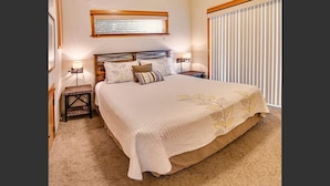 A bedroom with a large bed, two bedside tables, a nightstand, a window with blinds, and a wall-mounted lamp. - 2-story, 3 bedroom modern home with king and queen beds, living room and kitchen. (Ashland)
