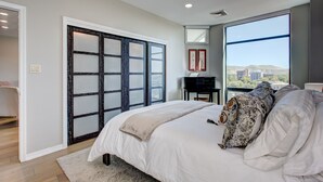 Un dormitorio con una cama grande, un escritorio y vistas a la ciudad a través de un ventanal.