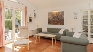 Een woonkamer met een bank, fauteuil en salontafel. Aan de muur hangt een schilderij, er is een kast met servies en een raam met gordijnen.
