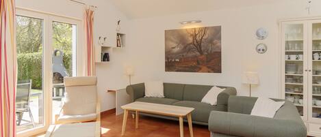 Un salon avec un canapé, un fauteuil et une table basse. Il y a un tableau au mur, un buffet rempli de vaisselle et une fenêtre avec des rideaux.
