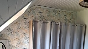 A room with a sloped ceiling, a large window, a grey curtain, a blue chair, and a patterned wallpaper.