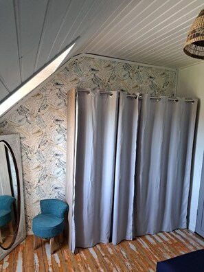 Ein Zimmer mit schrägem Dach, einem großen Fenster, einem grauen Vorhang, einem blauen Stuhl und einer Wand mit Muster.