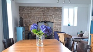 A dining area with a wooden table, chairs, a vase with flowers, a stone fireplace, and a window.